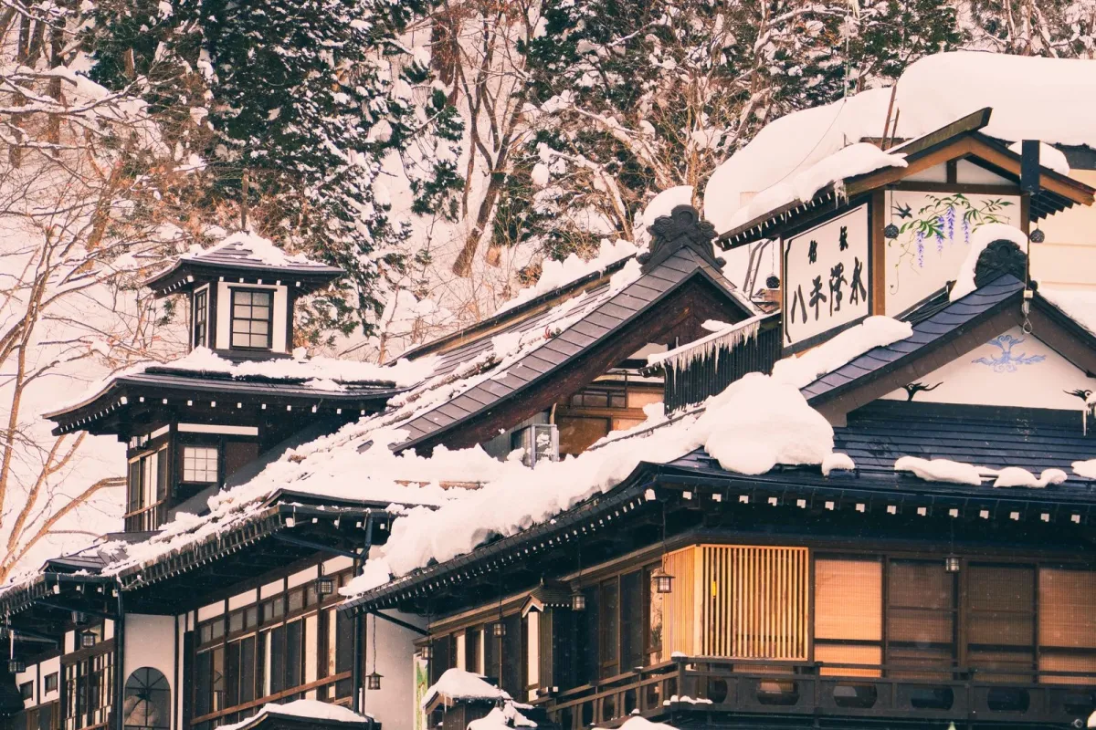 Traditional wooden ryokan nestled among autumn mountains with natural hot spring steam rising beside a stone garden and river.