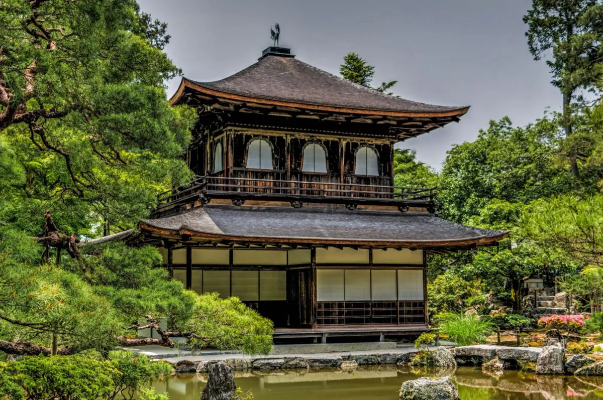 Zen monk in black robes sitting in meditation posture within minimalist temple room with natural light filtering through shoji screens.
