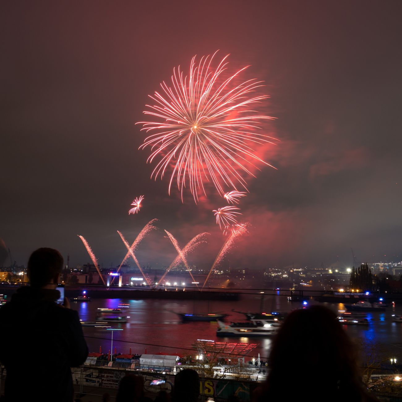 Colorful fireworks bursting over a Japanese riverside at dusk as crowds in yukata gather on blankets to watch the summer display.
