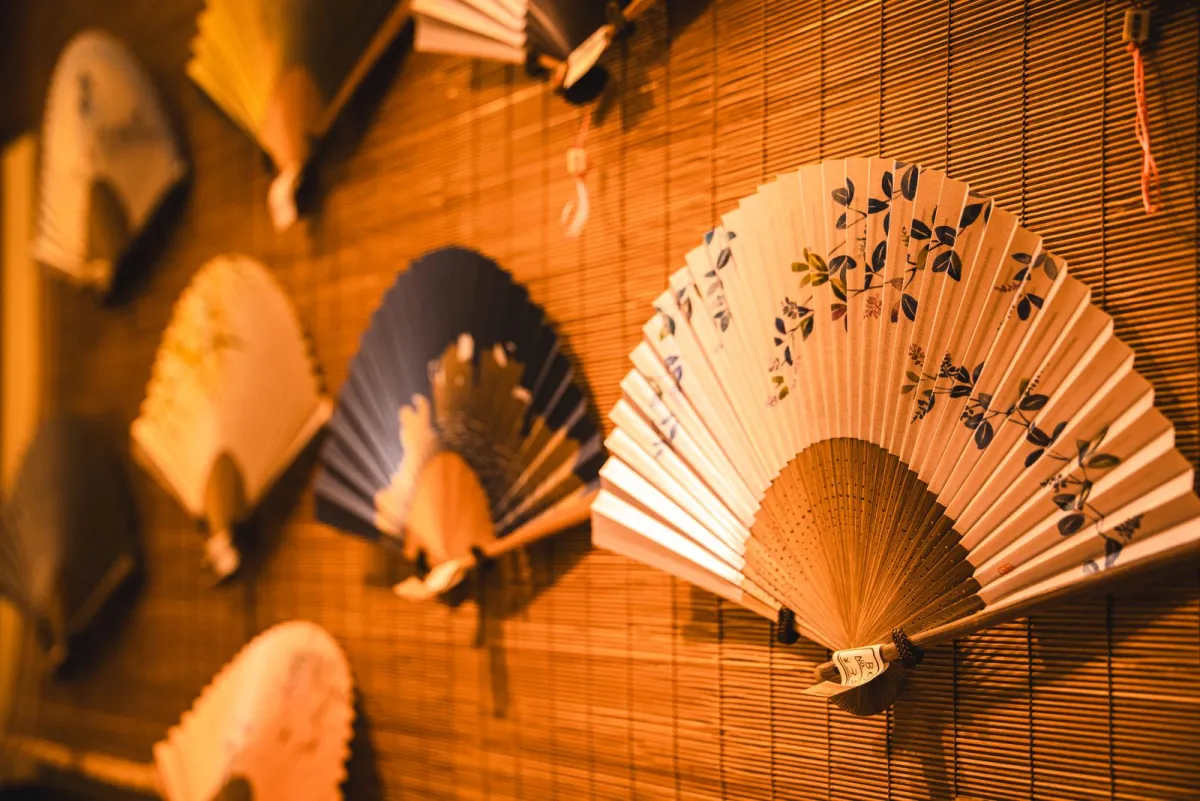 Craftsman's hands painting delicate cherry blossoms onto traditional Kyoto sensu fan ribs with fine bamboo brush.