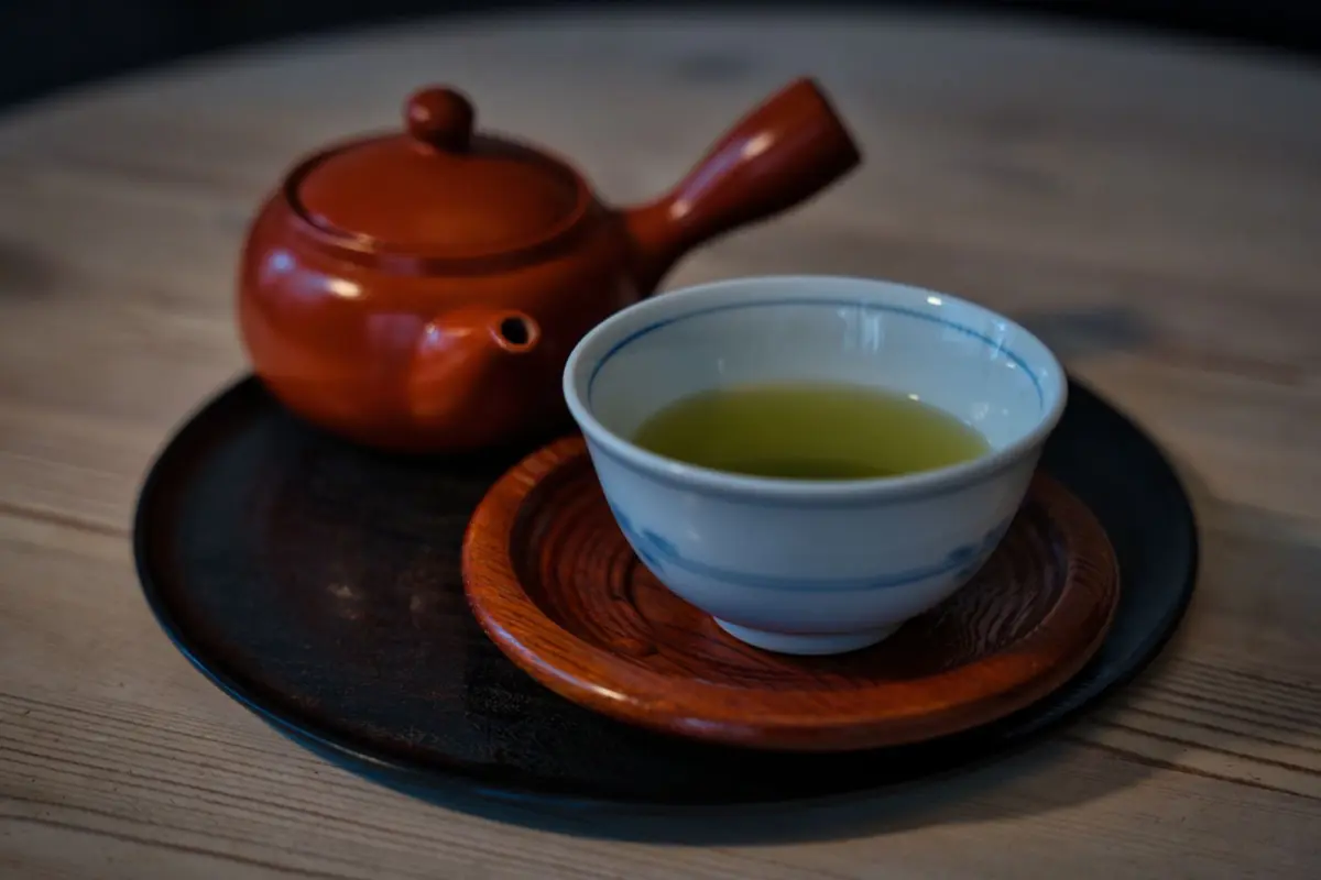 Traditional Tokoname red clay teapot with natural iron-rich surface beside green tea leaves on wooden table.