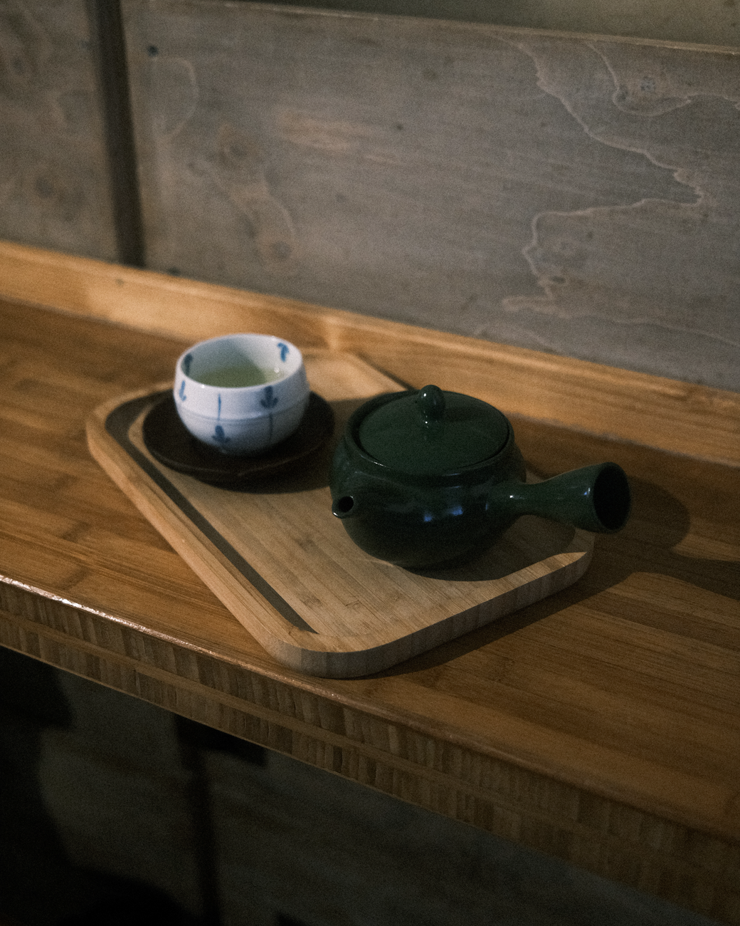 Japanese person pouring green tea from a ceramic kyusu teapot into a small handleless cup on a wooden table.